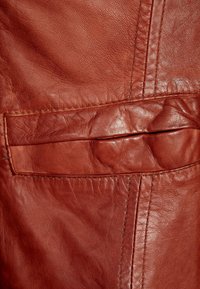 Hnědá kožená textura s hladkým povrchem a jemnými vráskami. Obsahuje kapsu s prošívaným obrysem pro detail.