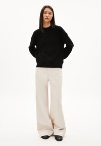 Schwarzer Strickpullover mit Rundhalskragen und überschnittenen Schultern, kombiniert mit beigen Culotte-Hosen. Die Schuhe sind schwarz und haben eine glatte Oberfläche.