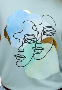 Lichtblauw t-shirt met abstracte lijntekening van twee gezichten, voorzien van kleuraccenten in groen en goud. Zachte stof met een relaxte pasvorm.