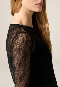 Top de encaje negro con mangas largas, que presenta patrones florales, un escote redondo y un cuerpo liso. La tela tiene una apariencia delicada y texturizada.