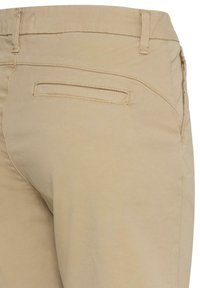 Pantalon beige montrant une poche arrière avec une fente horizontale, des passants de ceinture et une couture sur la couture latérale.