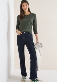 Haut vert à manches longues associé à un jean évasé bleu foncé. Le jean présente des coutures blanches contrastantes. Le mannequin tient un sweat gris et porte des baskets.