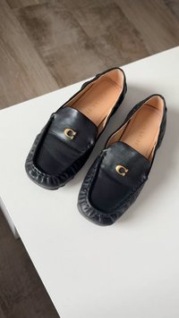 Sorte læder loafers med polstrede kanter, der har et guld logo detaljeret foran. Glat tekstur og rund tådesign.