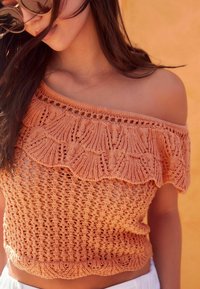 Ung kvinde med langt mørkt hår iført en orange off-shoulder strikket top og briller, mod en orange baggrund.
