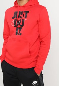 Sudadera roja con capucha, que presenta un gran logo negro de "JUST DO IT" y un bolsillo canguro. Combinada con pantalones negros que muestran el logo de Nike.