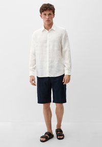 Weißes Leinenhemd mit langen Ärmeln, dunkelblaue Shorts und braune Sandalen, getragen von einer Person, die vor einem schlichten Hintergrund steht.