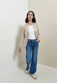 Blazer beige chiaro con una sottile texture, abbinato a una maglietta bianca e jeans a gamba larga blu. Sneakers multicolore completano l'outfit.