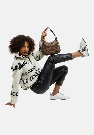 Frau mit lockigem Haar, die auf einer Hand und einem Fuß balanciert, trägt einen weißen bedruckten Fleece, schwarze Hose, weiße Turnschuhe und hält eine braune gemusterte Handtasche.