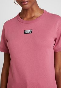 Růžové bavlněné tričko s kulatým výstřihem a krátkými rukávy, které na hrudi má černou obdélníkovou nášivku s logem Adidas.