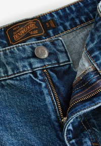 Jeans de denim albastru cu un buton și fermoar rezistente din argint. Prezintă un eticheta neagră cu text portocaliu, cusături întărite și o croială standard.