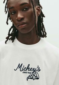 Persoană cu dreadlocks negre, purtând o cămașă albă cu textul "Mickey's Since 1928" și un grafic cu o felie de pizza, în text negru.