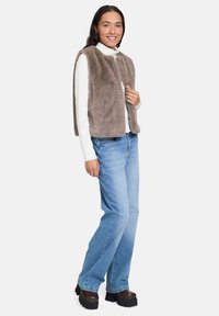 Junge Frau in einer taupefarbenen Kunstpelz-Weste, einem weißen Rollkragenpullover, blauen Jeans und klobigen braunen Plateau-Schuhen, lächelnd mit einer Hand auf der Weste.