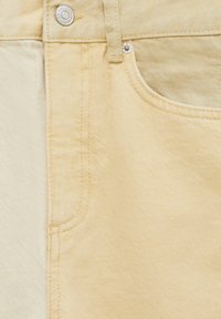 Gros plan sur un tissu en denim beige bicolore avec une poche avant, une passante de ceinture et des rivets en métal sur une paire de jeans.