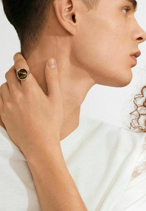 Close-up van het profiel van een jonge man met kort haar, gekleed in een wit shirt, en een hand met een grote donkere ring die op zijn nek rust.