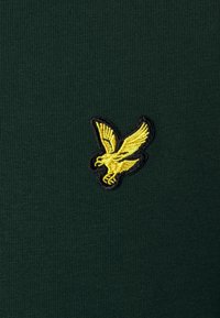 Zelené tričko s krátkým rukávem, na levé straně vyšívané žluté logo orla s černým obrysem. Hladká textura a přiléhavý střih.