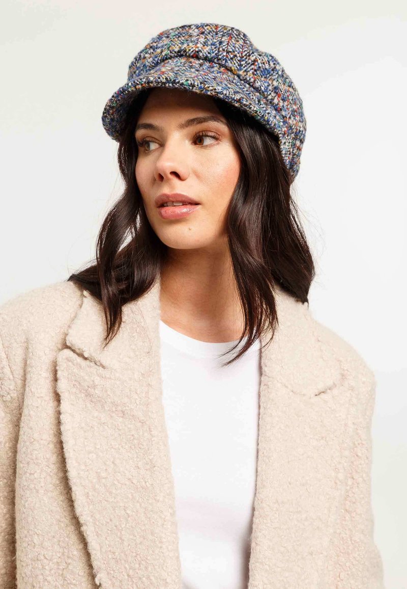 Vlnená čiapka s viacfarebným tweedovým vzorom, s širokým okrajom a textúrovaným povrchom. Nosená s béžovým kabátom cez biely top.