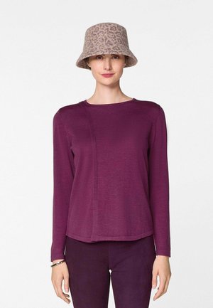 Femme portant un bob beige à imprimé léopard, un haut à manches longues violet et un pantalon violet assorti, debout devant un fond blanc.