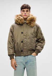 Młody mężczyzna w zielonej bomberce z dużym brązowym kapturem wyłożonym futrem, w zestawieniu z jasnoniebieskimi dżinsami, stojący na jednolitym tle.