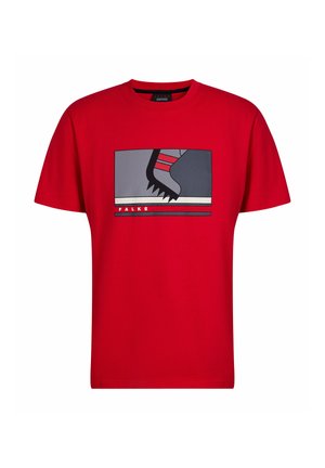 Lachende man die staat, gekleed in een rood grafisch T-shirt, zwarte shorts, zwarte sokken en zwarte sportschoenen tegen een effen witte achtergrond.