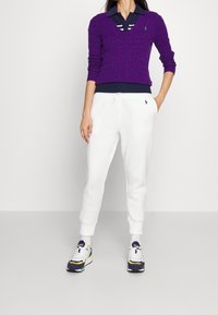 Pull en maille torsadée violet sur un polo bleu marine rayé, associé à un pantalon de jogging blanc et des chaussures de sport multicolores.