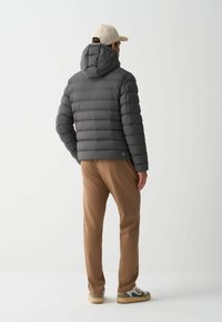 Grå pufferjakke med hætte, med quiltet tekstur. Båret med brune bukser og sneakers. Enkel design, uden synlig branding bagpå.