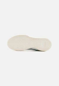 Scarpa con suola in gomma testurizzata, caratterizzata da scanalature intricate e dettagli con logo. Parte superiore grigia con accenti chiari, progettata per stabilità e presa.