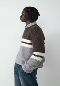 Flauschiger Strickpullover in Braun, Grau und Weiß mit Streifen; runder Ausschnitt; über einem karierten Hemd getragen; kombiniert mit blauen Jeans.