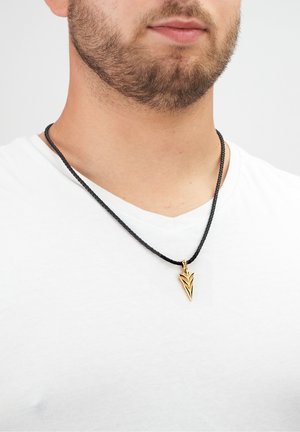 Schwarze geflochtene Halskette mit einem goldenen Anhänger in Form eines stilisierten Pfeils, getragen auf einem weißen Hemd.