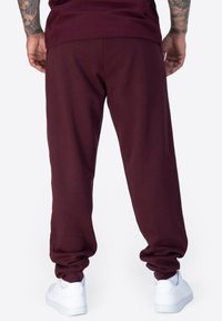 Bordeauxfarbene Sweatpants mit lässigem Schnitt, empfangenen Knöcheln und glatter Textur; kombiniert mit weißen Sneakers, die ein Logodetail präsentieren.