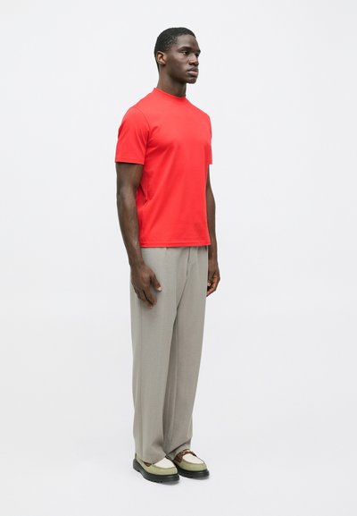 Άντρας που στέκεται φορώντας ένα φωτεινό κόκκινο κοντομάνικο πουκάμισο, ανοιχτό γκρι χαλαρούς παντελόνες και δίχρωμα loafers σε λευκό φόντο.