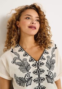 Blusa de lino blanca bordada con patrones florales negros, mangas cortas y un escote en V con un intrincado detallado a lo largo de los bordes.