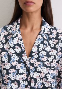 Mujer con una parte superior de pijama oscura de flores, con flores en tonos rosa claro, blanco y azul, y un bolsillo en el pecho con detalles de ribete negro.