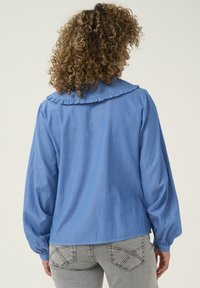 Blusa blu a maniche lunghe con colletto arricciato e polsini svasati, realizzata in tessuto leggero. Indossata con jeans grigi chiari caratterizzati da cuciture sui taschini posteriori.
