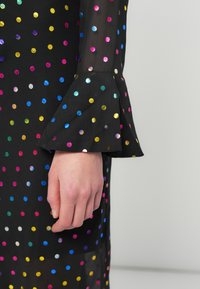 Μαύρο φόρεμα με πολύχρωμες μεταλλικές βούλες. Διαθέτει μανίκι καμπάνα στον καρπό, αναδεικνύοντας μια λεία υφή και ζωντανά χρώματα.