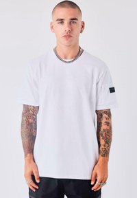 Joven con cabello corto y tatuajes en ambos brazos, lleva una camiseta blanca, un collar de cadena de plata y varios anillos, todo sobre un fondo sencillo.