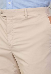 Beige bomullsbukser med flat front, sidelommer og en enkelt knappelukking i livet, vist mot en lys blå skjorte.