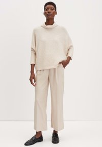 Maglione lavorato a maglia di un beige chiaro con un collare alto, abbinato a pantaloni su misura color crema e mocassini neri. Texture liscia e vestibilità comoda.