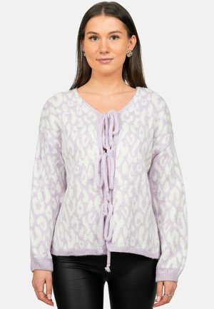Woman wearing a lavender and white patterned knitted cardigan with three front tie closures and black pants.