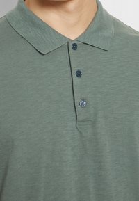 Gros plan sur une personne portant un polo vert avec un col et trois boutons foncés fermés.