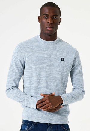 Man met een donkere huid die een lichtgrijze gebreide trui met een klein borstlogo draagt, staande met de handen voor zich in elkaar geklapt, tegen een effen witte achtergrond.