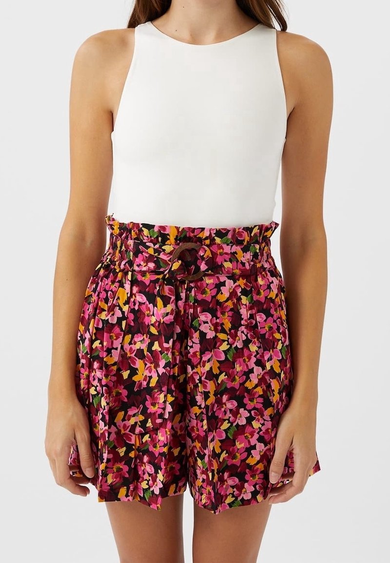 Femme portant un short taille haute avec un imprimé floral rose, orange et jaune, et un haut blanc sans manches avec un col rond.