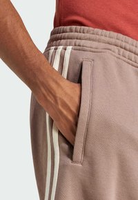 Pantalon de survêtement marron clair avec des bandes blanches sur les côtés, doté d'une poche latérale et d'une taille côtelée. Tissu en coton doux avec une coupe décontractée.