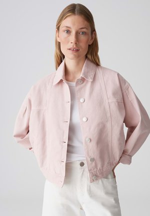 Donna con capelli biondi lisci che indossa una giacca rosa chiaro abbottonata sopra una maglietta bianca e pantaloni bianchi, in piedi con le mani sui fianchi.