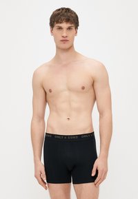 Černé bavlněné boxerky s přiléhavým střihem, s páskem se zlatým logem a hladkou texturou. Jednobarevné pozadí.