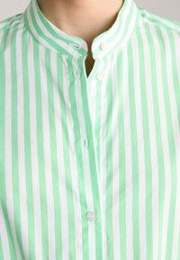 Zielono-biała koszula w paski z kołnierzem stójką, zapinana na guziki, wykonana z miękkiej bawełny. Paski mają jednakową szerokość.