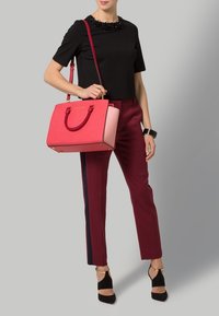Borsa strutturata rossa e rosa con doppi manici e dettagli dorati, indossata con un top nero e pantaloni borgogna con una striscia laterale.