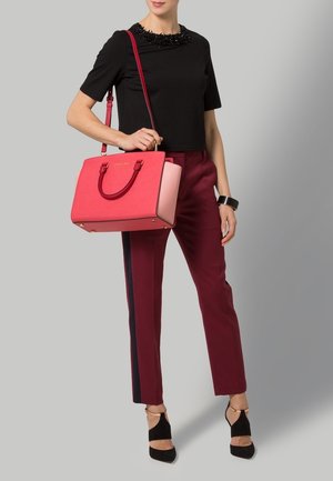 Sac à main structuré rouge et rose avec double poignée et accents dorés, porté avec un haut noir et un pantalon bordeaux avec une bande latérale.