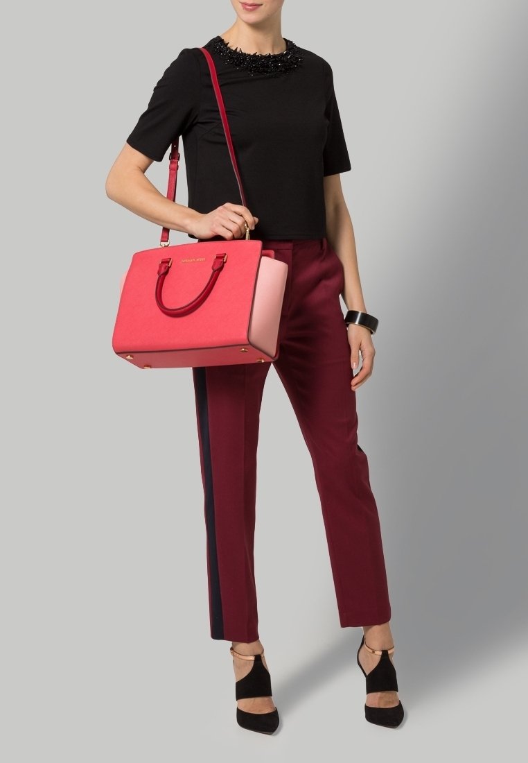 Borsa strutturata rossa e rosa con doppi manici e dettagli dorati, indossata con un top nero e pantaloni borgogna con una striscia laterale.
