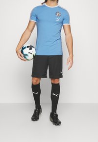 Mužský fotbalista v světle modrém dresu Manchester City, černých šortkách, černých ponožkách a kopačkách, drží míč Puma u svého boku.