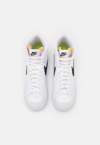 Białe trampki typu high-top z czarnym logo Swoosh, wyściełanym kołnierzem oraz jaskrawymi zielonymi i żółtymi akcentami na języku i wkładce. Teksturowana gumowa podeszwa.